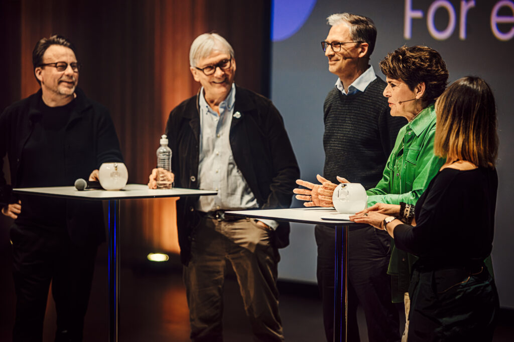 Fem personer står på scen i ett engagerat samtal där de riktar sig mot varandra med ett fokuserat uttryck och den övergripande känslan är lugn och närvarande.