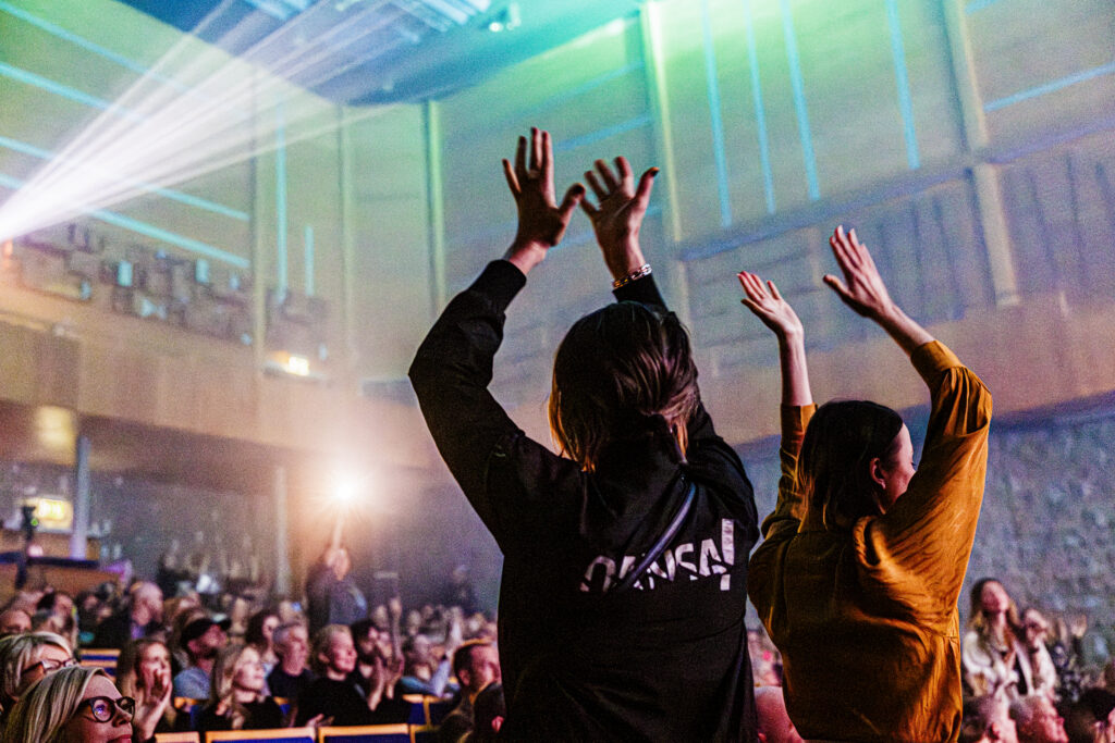 Människor reser sig och applåderar i en konsertlokal fylld av färgade ljus där den glada känslan sprider sig mellan publiken och scenen.