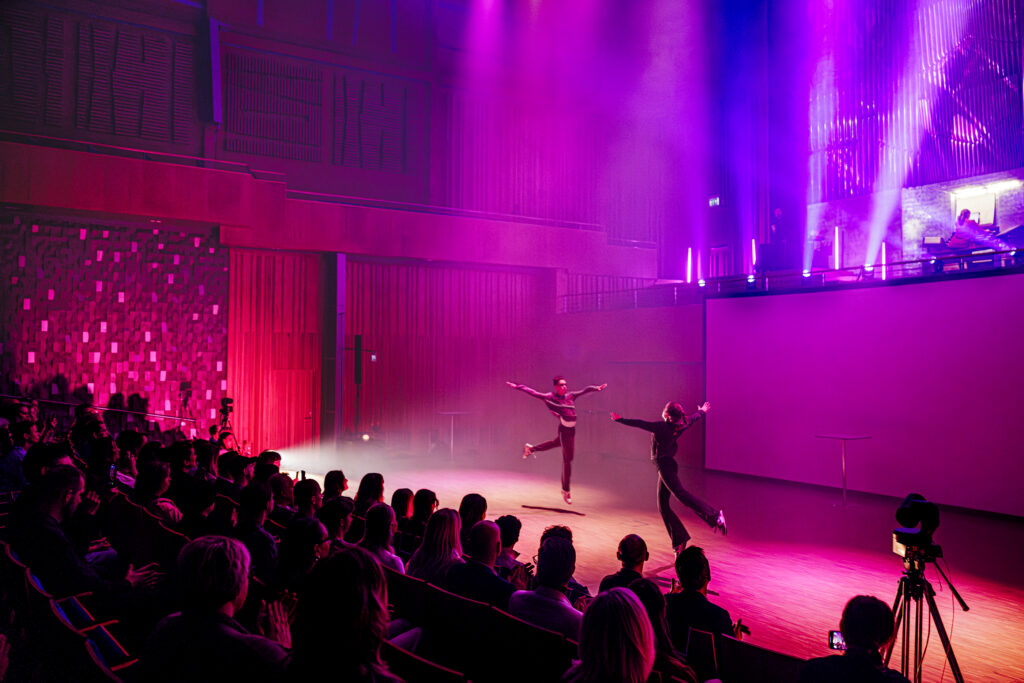 Två personer dansar på scen i färgade scenljus medan publiken ser på och den glada känslan i lokalen förstärks av rörelserna.