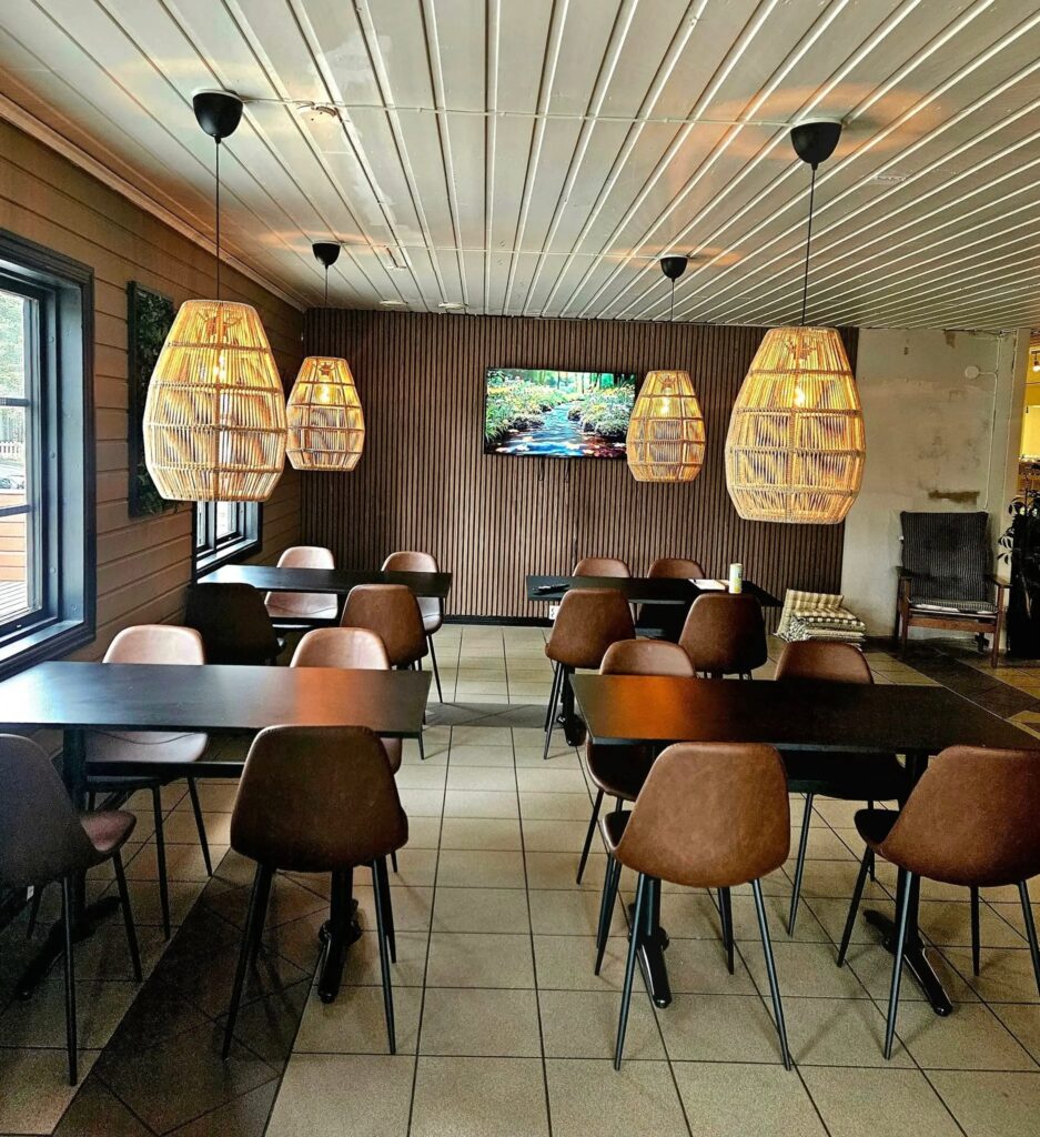 Kafémiljö i varma och vilsamma toner med bord och stolar utspridda i rummet. Stora ljusa armaturer hänger ovanför och ger ett mjukt sken.