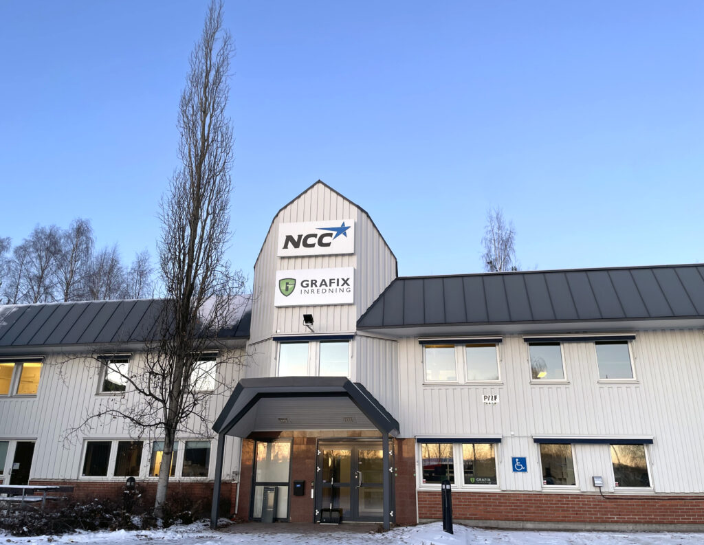 Exteriör av vit plåtfasad med tydlig entré i mjukt vinterljus. Snö täcker marken och på fasaden syns skyltar för Grafix inredning och NCC.
