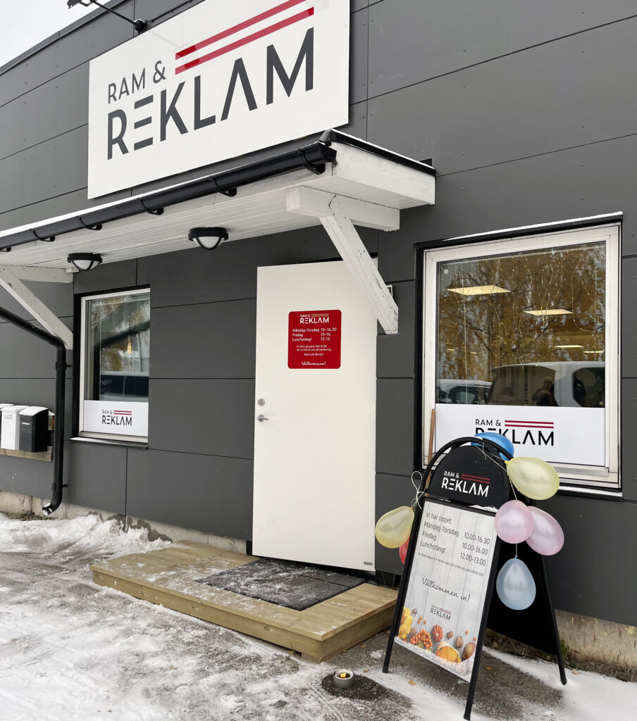 En entré med trästeg leder fram till en grå fasad och en vit entrédörr där en röd dekal med företagsnamnet Ram och reklam sitter centrerad. Rena linjer och stilla ljus ger ett välkomnande intryck.