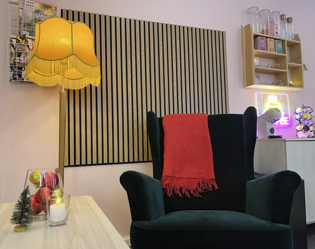 Mysig sittplats med mörkgrön fåtölj, röd pläd och gul golvlampa. På väggen bakom sitter träpanel och små hyllor med glas och dekorationer.