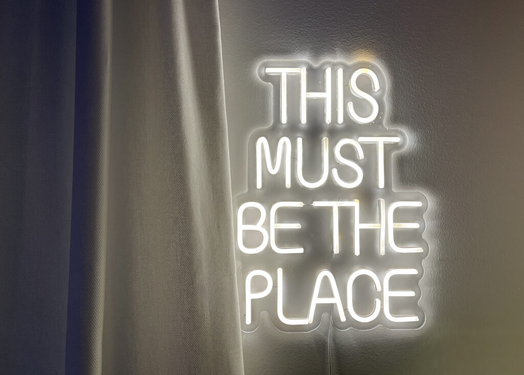 Vit neonskylt på ljus vägg med texten “THIS MUST BE THE PLACE”. Skylten lyser mjukt bredvid en gardin i grå toner.