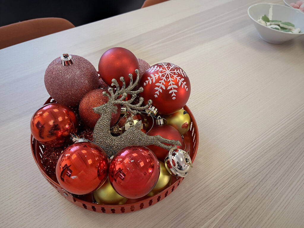Skål fylld med julkulor i rött och guld samt en glittrande ren. Skålen står på ett ljust bord med suddiga stolar i bakgrunden.
