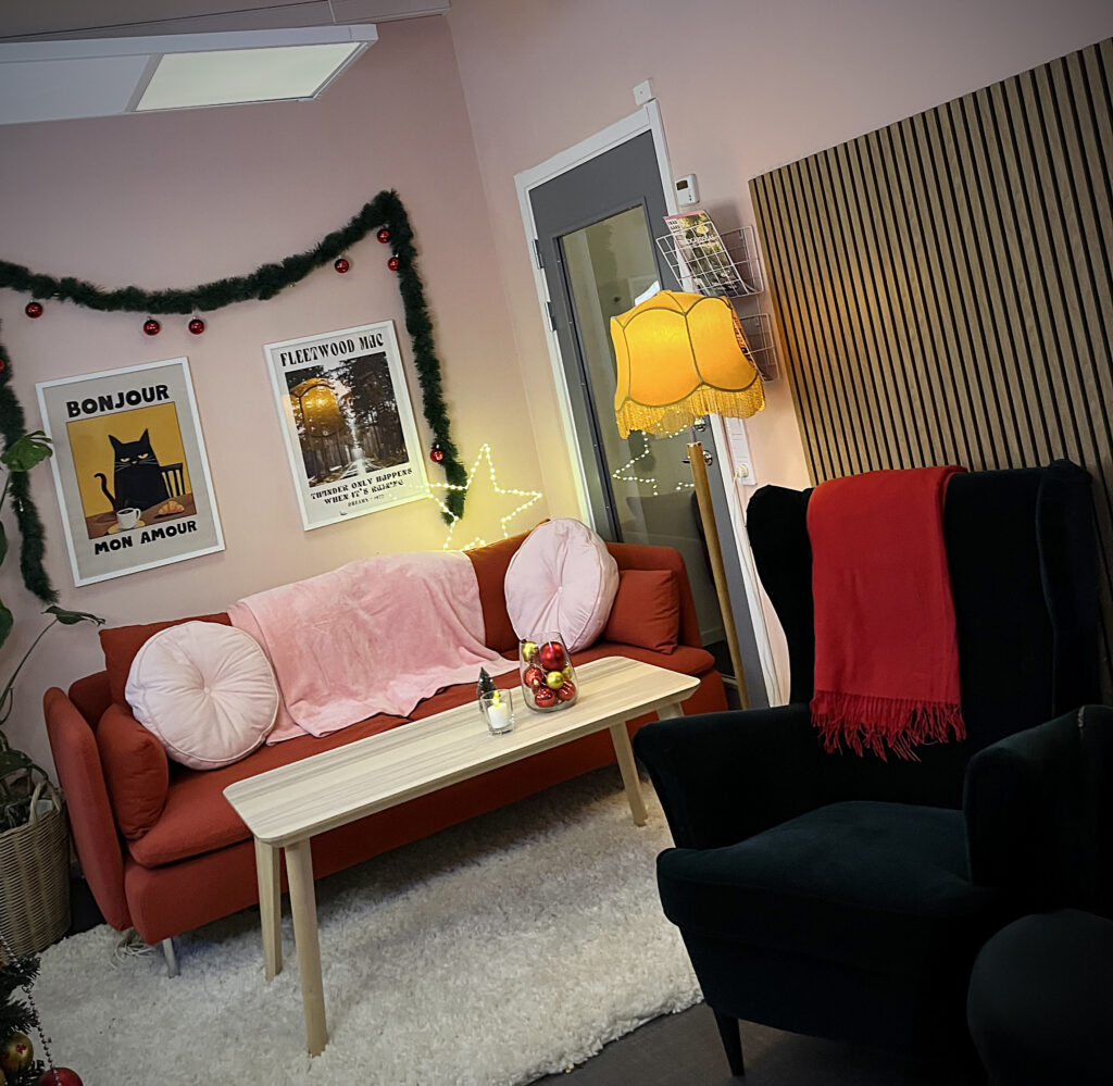 Sned vy över soffgrupp med rosa och röda textilier, litet bord och fåtölj. En gul golvlampa och juldekorationer ger rummet mjukt ljus.