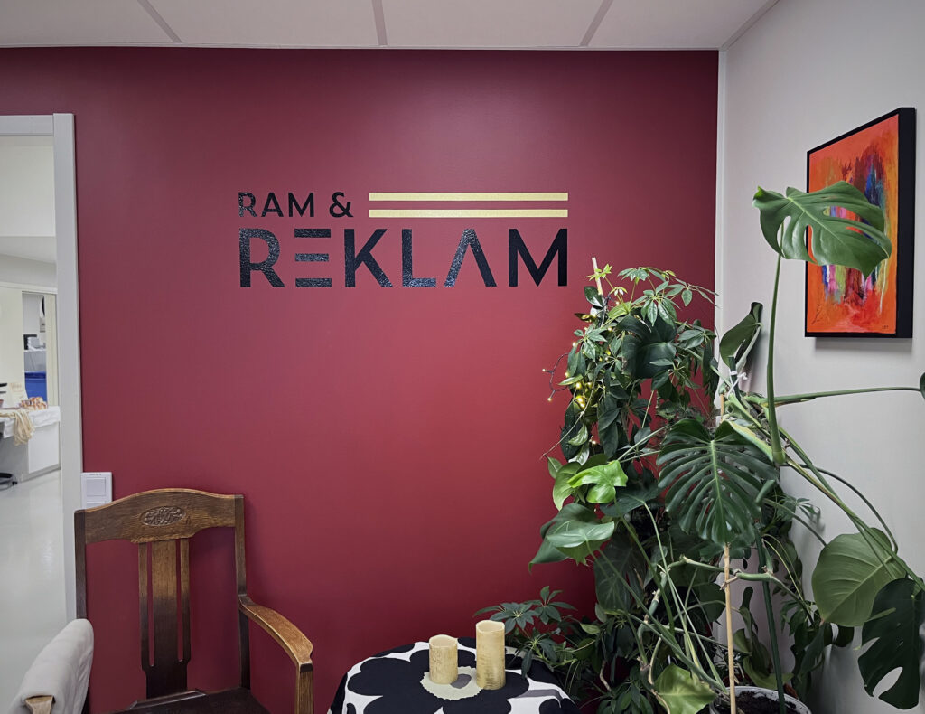 En fondvägg i djup kulör bär företagets svarta logotyp Ram och reklam. I förgrunden syns en stor växt med gröna blad som mjukar upp helheten.