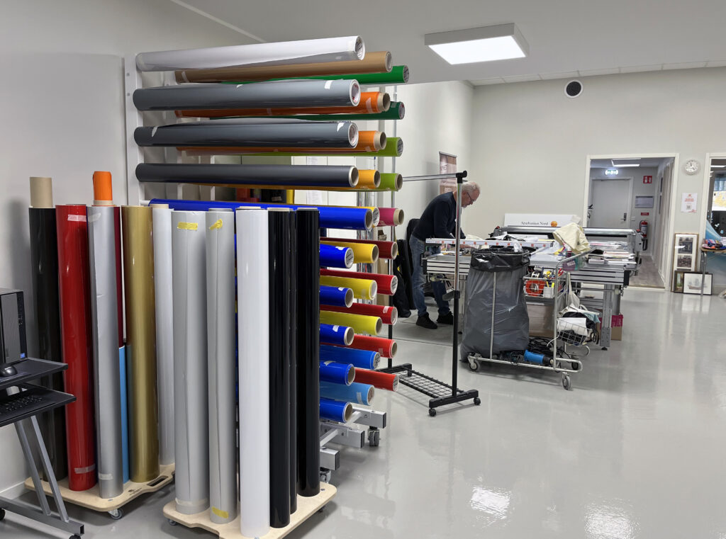 En rymlig och ljus produktionsyta i ett reklamföretag där rullar med dekaler i olika kulörer hänger på ett ställ och en man arbetar vid ett arbetsbord.