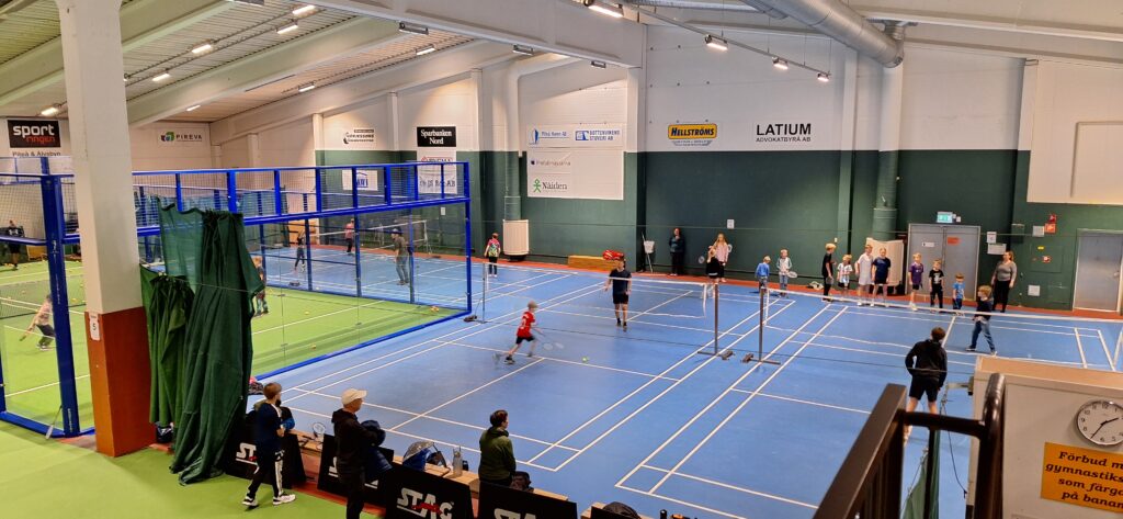 En stor inomhushall med flera badmintonbanor. Barn och vuxna tränar samtidigt medan ledare och föräldrar står längs kanten och tittar på.