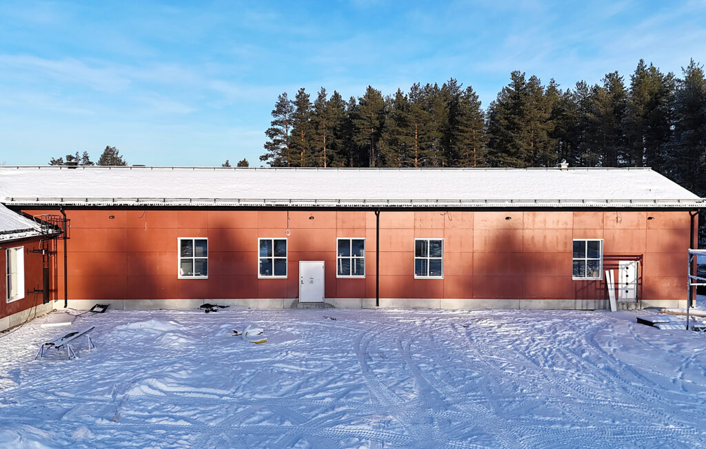 En lång röd fasad med flera fönster och snötäckt tak. Framför byggnaden ligger en stor vit gårdsplan med spår i snön.