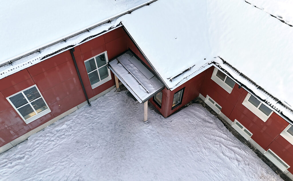 Flygbild ovanifrån av en röd byggnad i snö. Ett mindre entrétak syns i vinkeln mellan huskropparna.