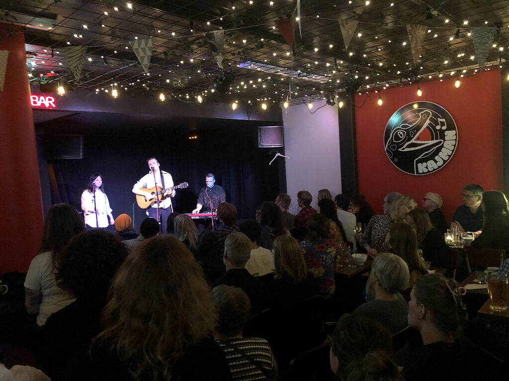 En liten konsertscen på en bar med tre musiker under varm belysning. Publiken sitter tätt vid bord och lyssnar, med glas och ljusslingor runtom.