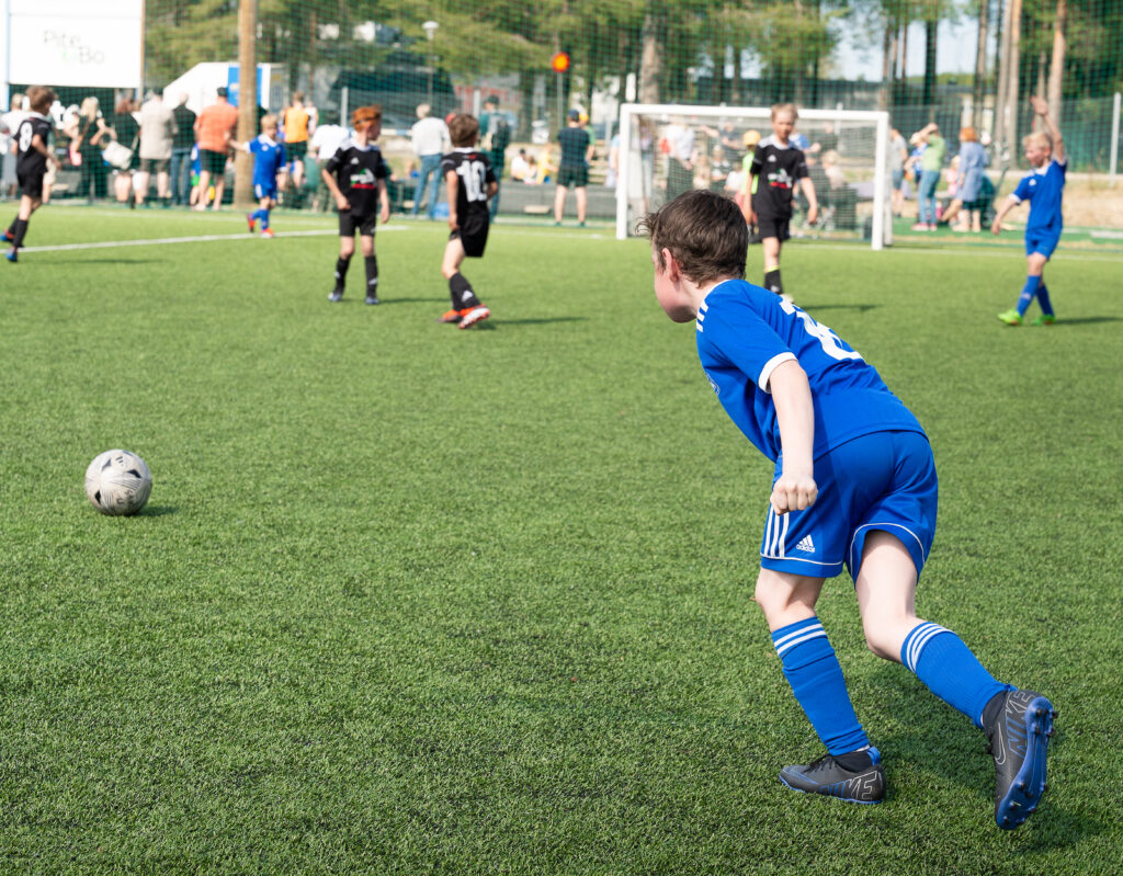 Ett barn i blå fotbollströja springer mot bollen på en grön konstgräsplan. Andra barn spelar i bakgrunden med mål och publik bakom.