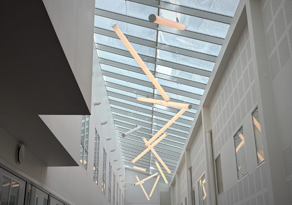 Ljust atrium med glastak där snön syns ovanifrån. Avlånga pendellampor hänger i olika vinklar och skapar ett grafiskt, luftigt rum.