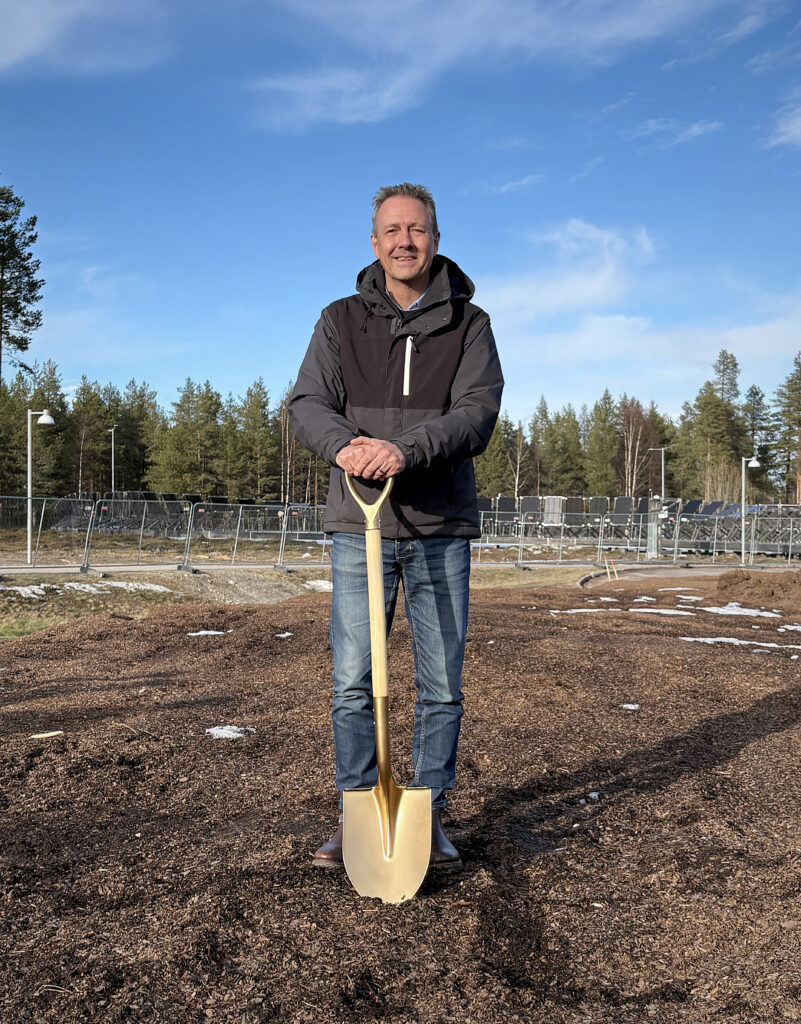 En man i mörk jacka och jeans står på grusig mark med en gyllene spade framför sig. Bakom syns stängsel, solpaneler och skog under klarblå himmel.