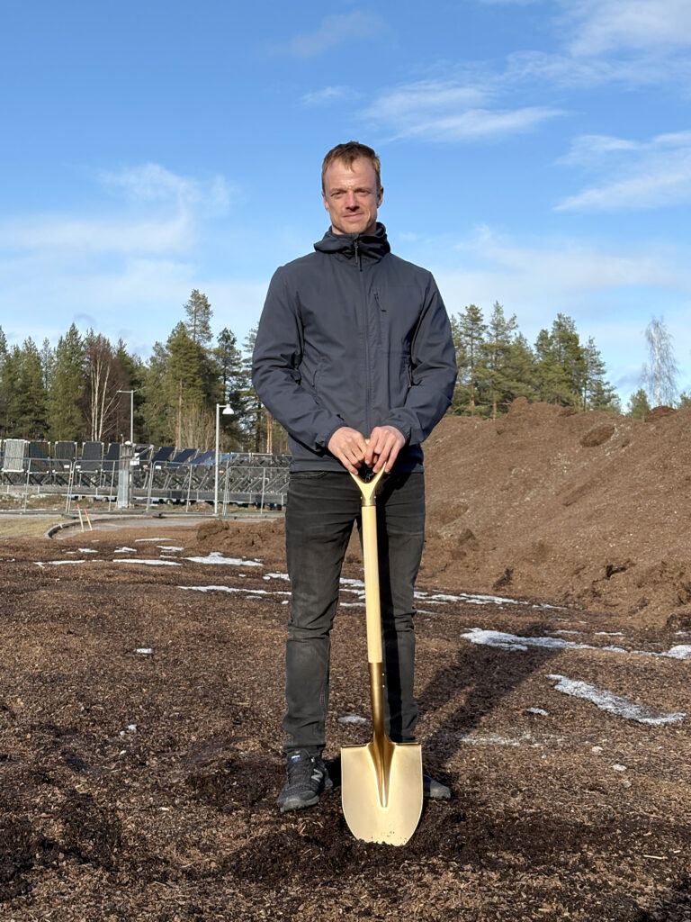 En man i mörk jacka och svarta jeans står stadigt med händerna på en gyllene spade. I bakgrunden syns jordhögar, stängsel och gles skog.