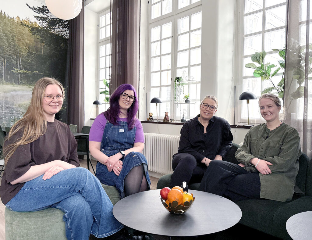 Fyra kvinnor sitter i fåtöljer runt ett runt bord med frukt. De ler mot kameran i ett ljust rum med stora fönster, växter och en skogsvy i bakgrunden.