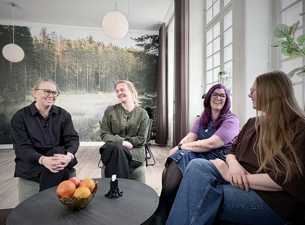 Fyra kvinnor sitter i en halvcirkel och samtalar. De skrattar och vänder sig mot varandra i ett ljust rum, med en stor väggbild av skog och sjö bakom.