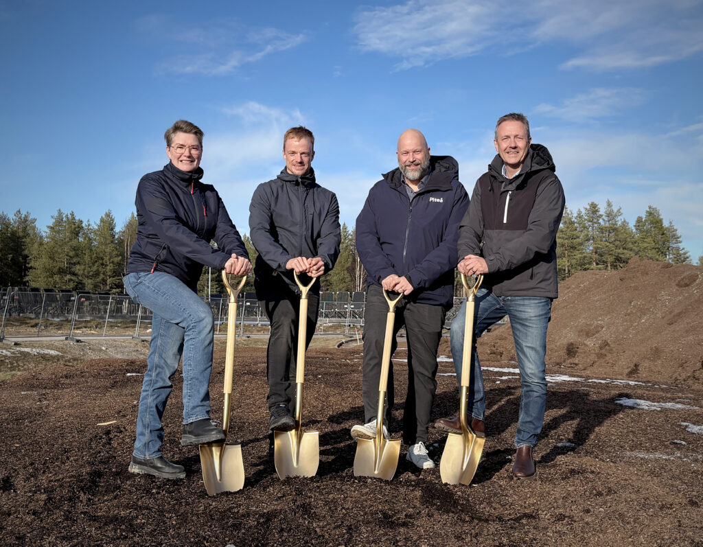 Fyra personer står sida vid sida med varsin gyllene spade, en fot placerad på bladet. De ler mot kameran, med byggområde och skog bakom.