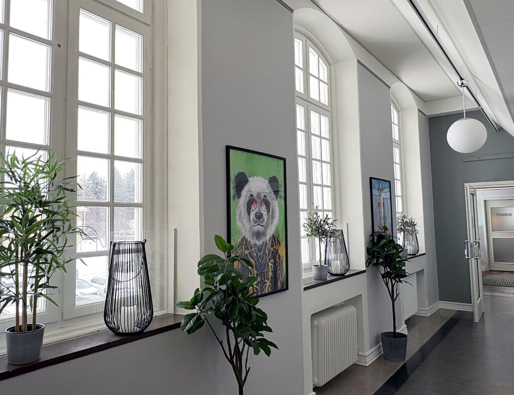 Lång korridor med höga fönster och växter på fönsterbrädorna. På väggarna hänger färgstarka djurporträtt, bland annat en panda i solglasögon.
