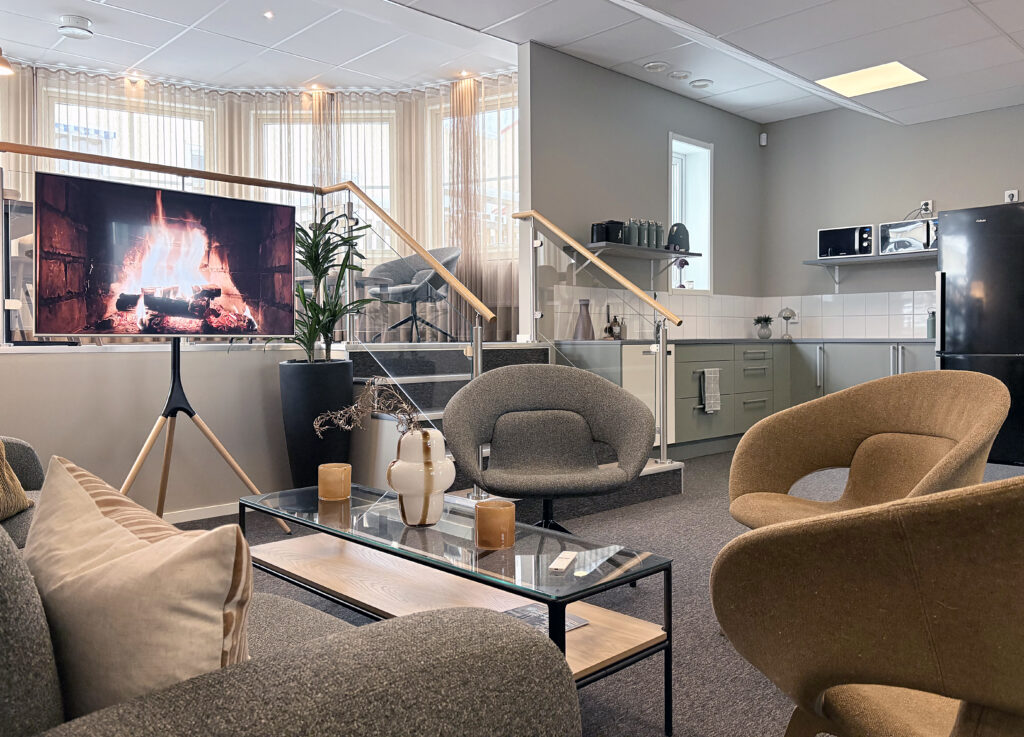 Ljus kontorslounge med grå soffa och fåtöljer i jordtoner. På väggen syns logotypen placerum. En tv visar en sprakande brasa.