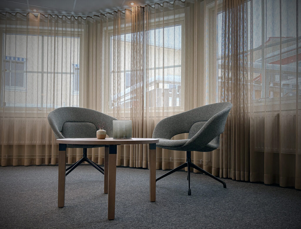 Ljus sittplats med två grå fåtöljer och ett runt träbord. Bakom hänger tunna gardiner framför stora fönster som släpper in mjukt dagsljus.