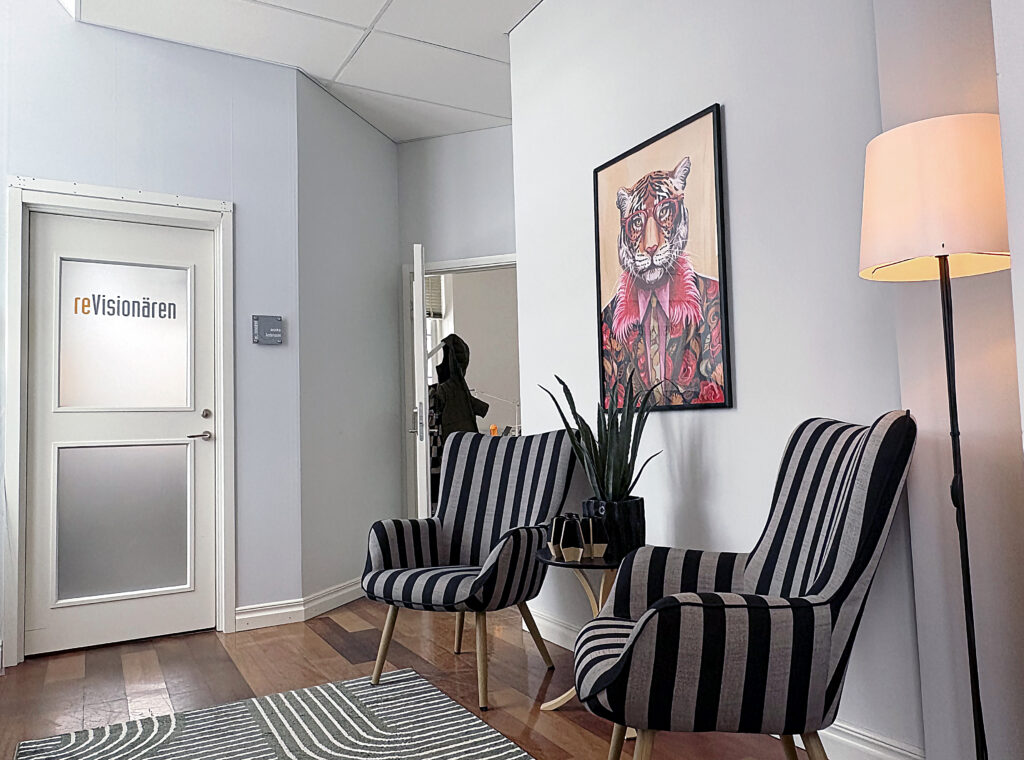 Två randiga fåtöljer står bredvid ett litet bord med växt och ljus. På väggen hänger en tavla med en tiger i glasögon, i ett lugnt hörn.