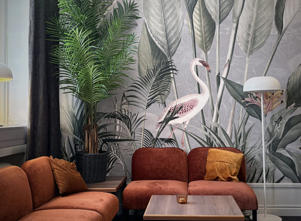 Rostfärgade soffor och gröna växter står framför en stor väggbild med flamingo och blad. Små bord och lampor skapar en varm loungemiljö.