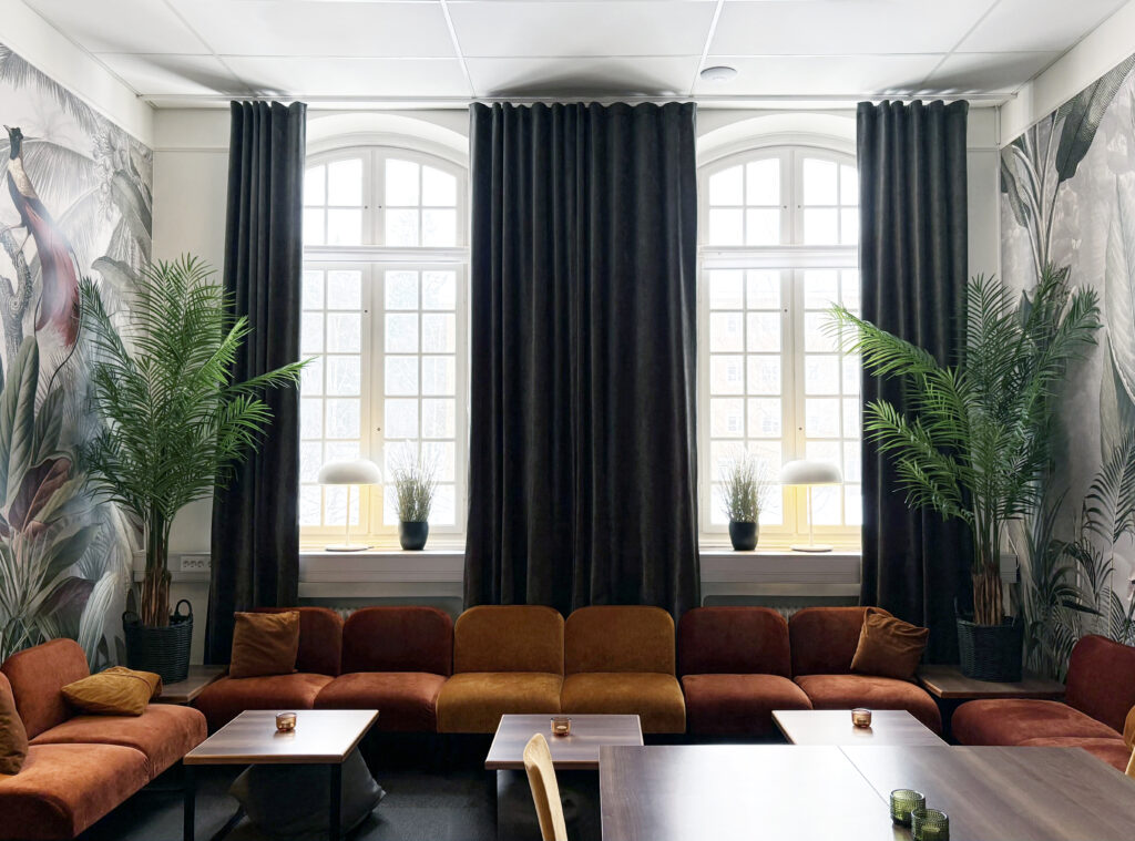 Mjuk soffgrupp i varma toner står framför höga fönster med mörka gardiner. Växter och små bord skapar en lugn loungeyta.