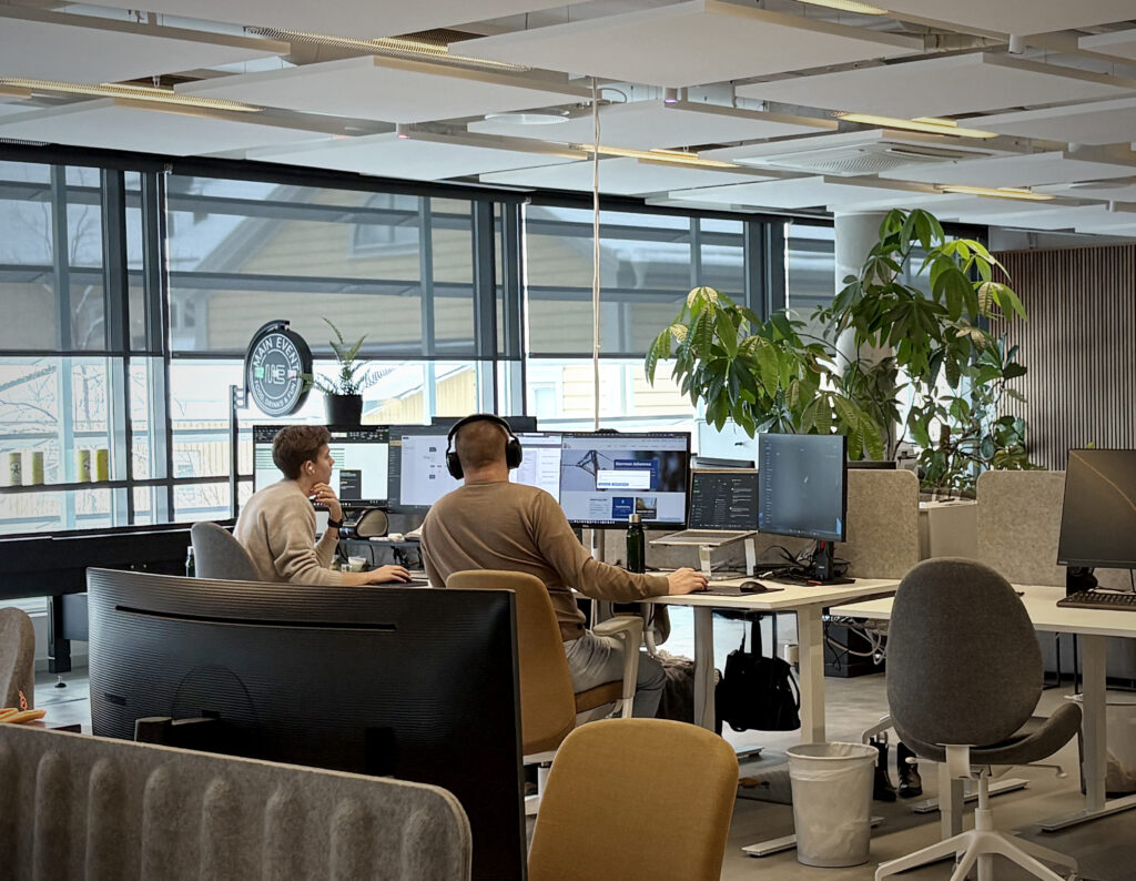 Öppet kontorslandskap med skrivbord i rader. Två personer arbetar vid skärmar, omgivna av växter och mjuka akustikpaneler.