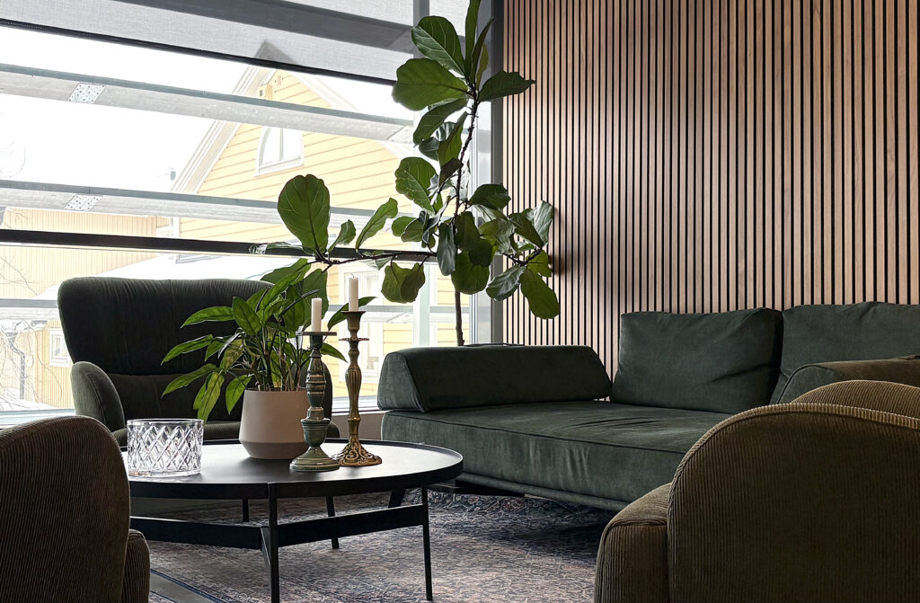 Loungehörna med gröna soffor, fåtöljer och runt bord. Stora växter och fönsterparti ger rummet ljus och en lugn, inbjudande känsla.