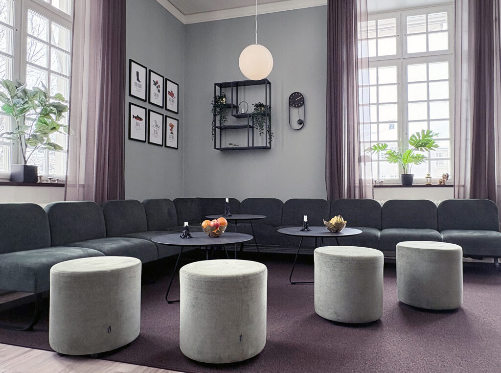 Stort sällskapsrum med soffor längs väggarna och flera runda sittpuffar. Bord med frukt och ljus står på en mörk matta.