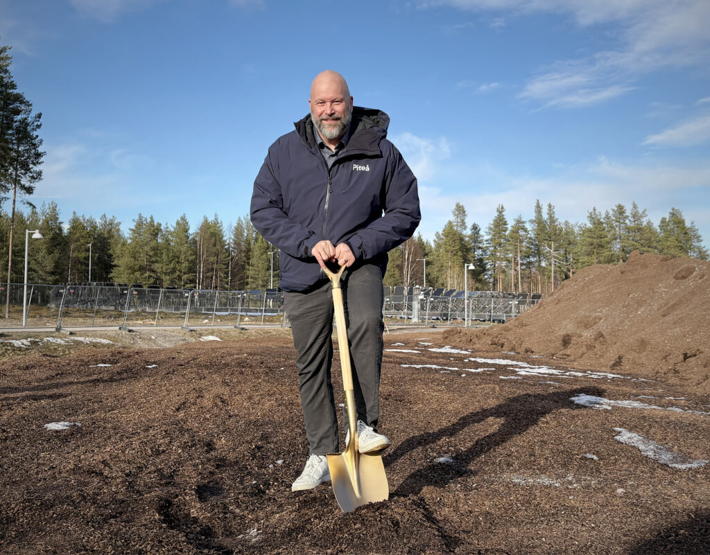 En man i mörk jacka med texten “Piteå” lutar sig mot en gyllene spade. Han ler lätt, med jordhögar, stängsel och skog i bakgrunden.