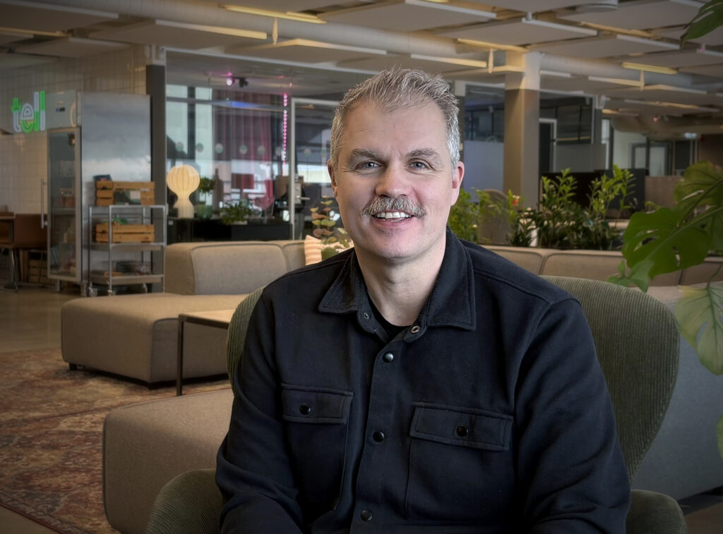 Närbild av samma man sittande i en grön fåtölj. Bakom syns öppet kontor med växter och mjuka former i en varm arbetsmiljö.