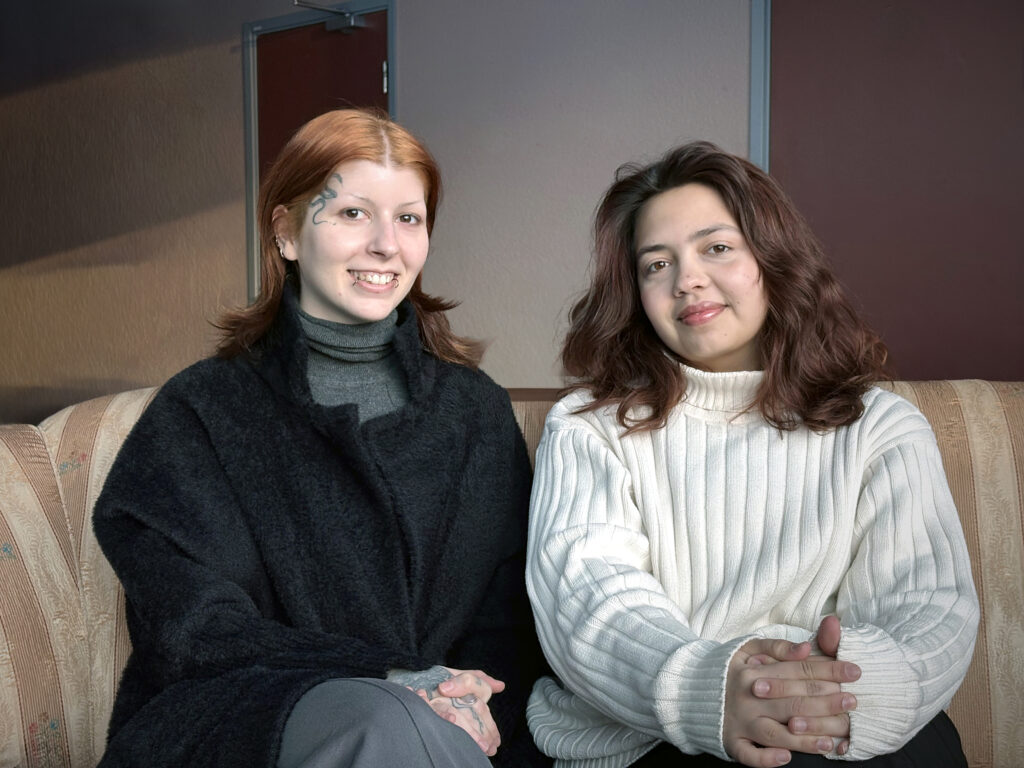 Två kvinnor sitter nära varandra i en soffa och ser mot kameran. Den ena bär svart kappa och polotröja, den andra en ljus stickad tröja.