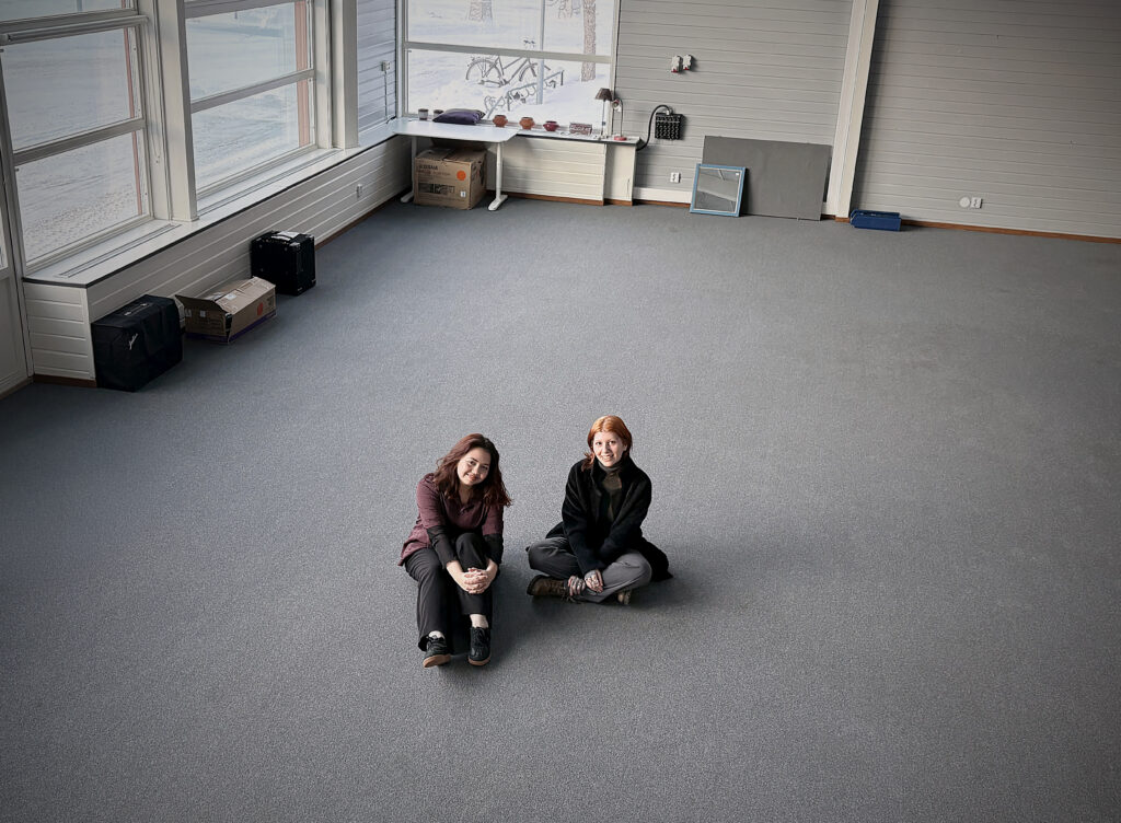 Två kvinnor sitter på golvet i ett stort, ljust rum med grå matta och stora fönster. De ler mot kameran. Utanför syns snö och vinterlandskap.