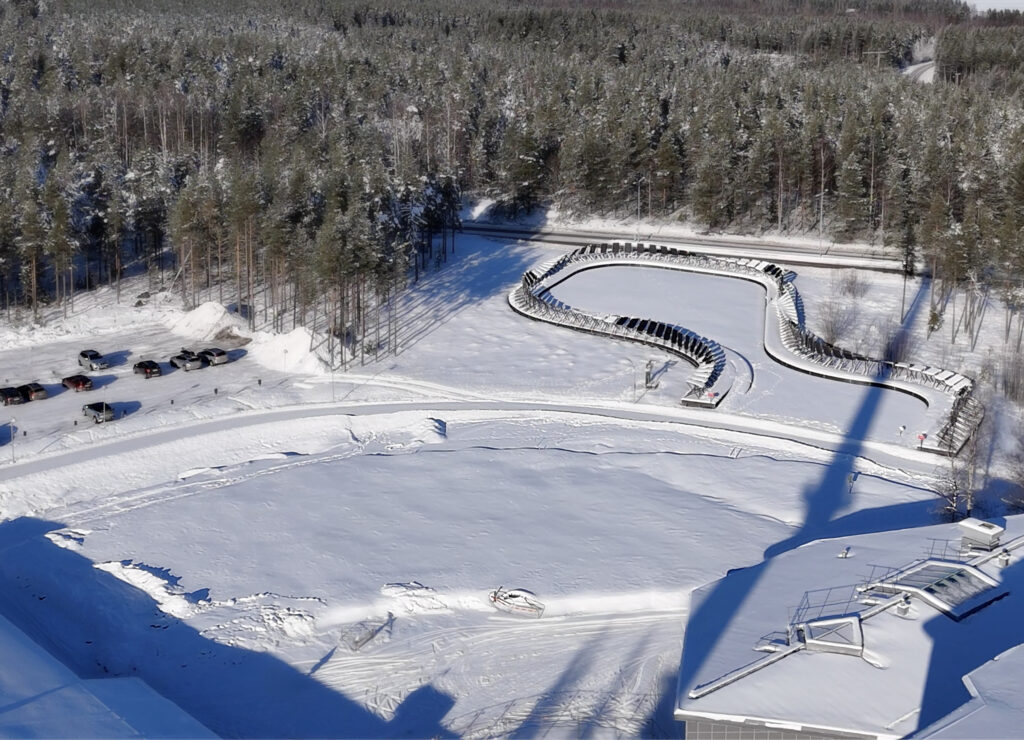 Snötäckt område sett uppifrån där marken förbereds för byggnation. En slingrande konstruktion löper genom området. Bilar står parkerade vid skogskanten och en väg passerar bakom.