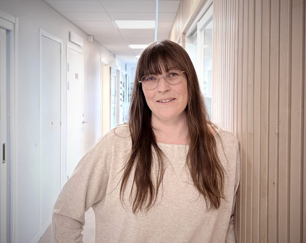 En kvinna med långt brunt hår och glasögon står i en ljus kontorskorridor. Hon ler lätt, bär en beige tröja och lutar sig mot en träpanel.