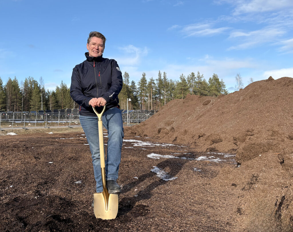 En kvinna i mörk jacka och jeans står med ena foten på en gyllene spade. Hon ler mot kameran, med jordhögar och skog bakom sig.