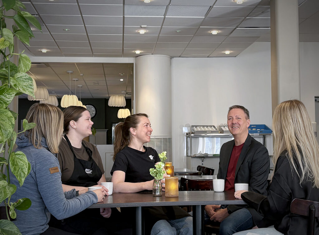 Fem personer sitter runt ett bord på ett café, ler och samtalar. Kaffekoppar, levande ljus och en grön växt skapar en varm, avslappnad miljö.