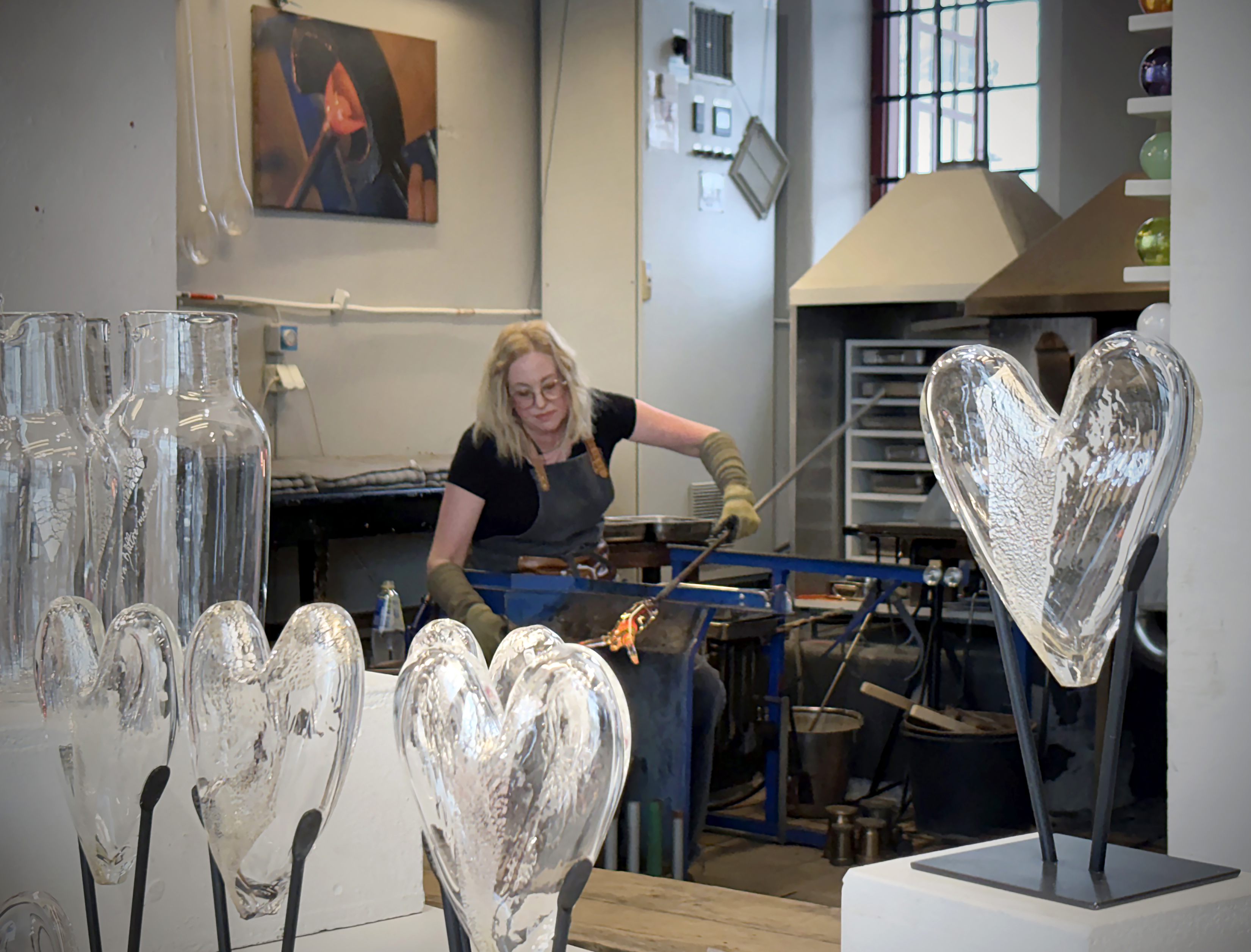 Kvinna arbetar med glas vid arbetsbänk. Hon formar glödande glas med verktyg. Färdiga glasfigurer står i förgrunden och gnistrar.