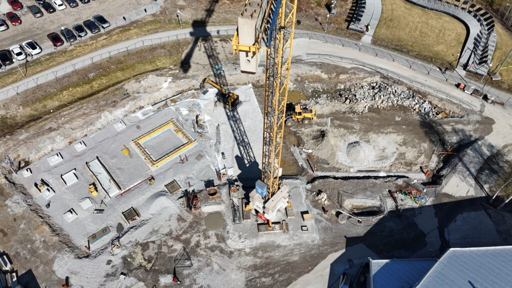 Flygbild över en byggarbetsplats där en tornkran reser sig mitt i området. Grusbäddar, gjutna fundament och arbetsmaskiner syns intill gångvägar och parkerade bilar.