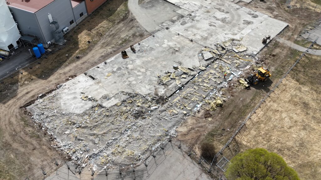 Drönarbild över rivningsområde där stora betongplattor brutits upp. En gul lastmaskin arbetar bland grus, damm och spridda isoleringsrester.