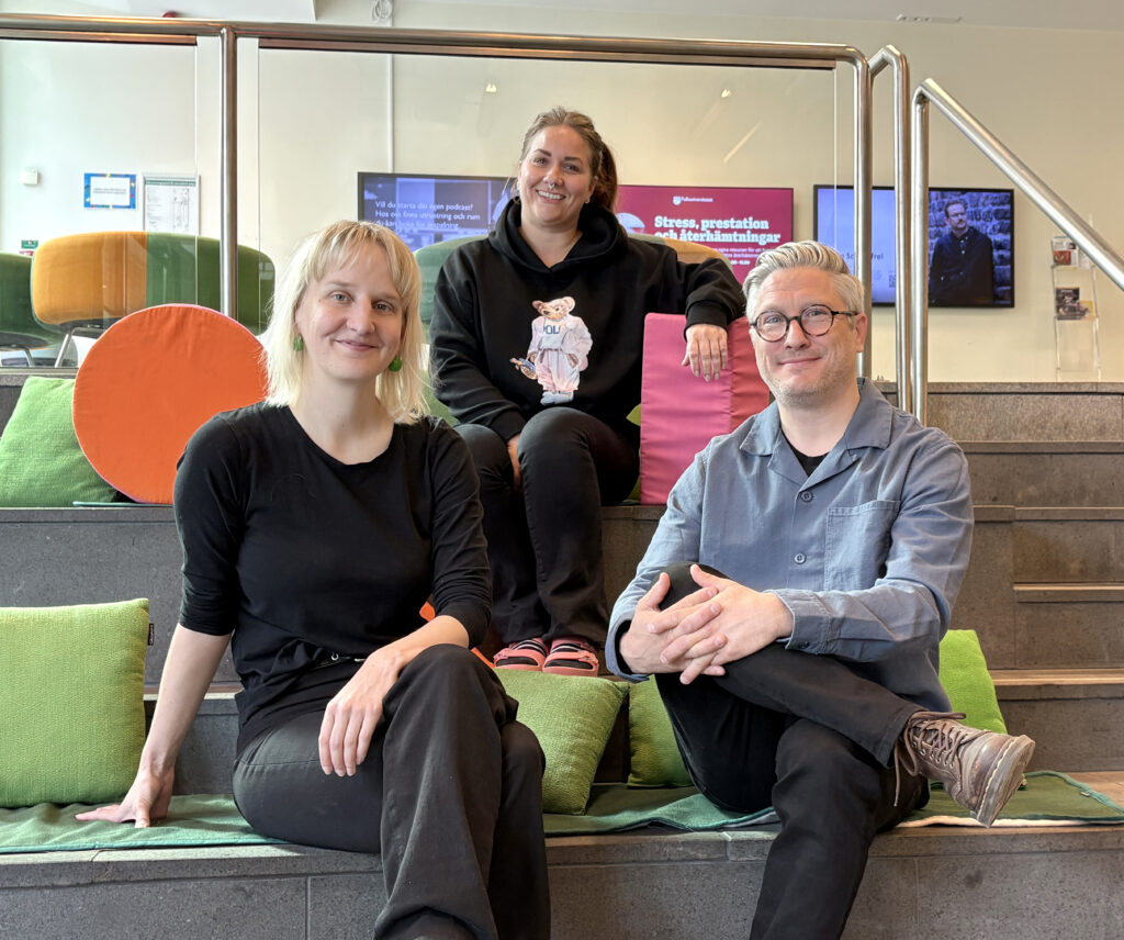 Tre personer sitter på mjuka kuddar i en färgstark samlingsyta med trappsteg, glasräcken och digitala skärmar i bakgrunden. Alla ler mot kameran.