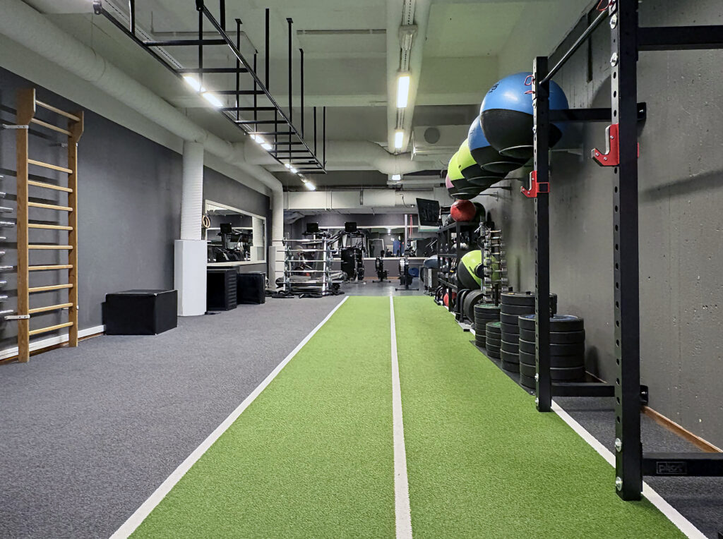 Lång grön löpbana för funktionell träning löper genom gymdel med medicinbollar, vikter och klätterställning.