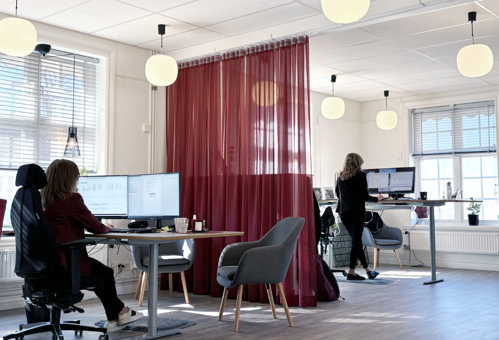 Ljust kontorslandskap med stora fönster, runda taklampor och vinröda textilskärmar mellan arbetsplatser där två personer arbetar vid höj- och sänkbara skrivbord.