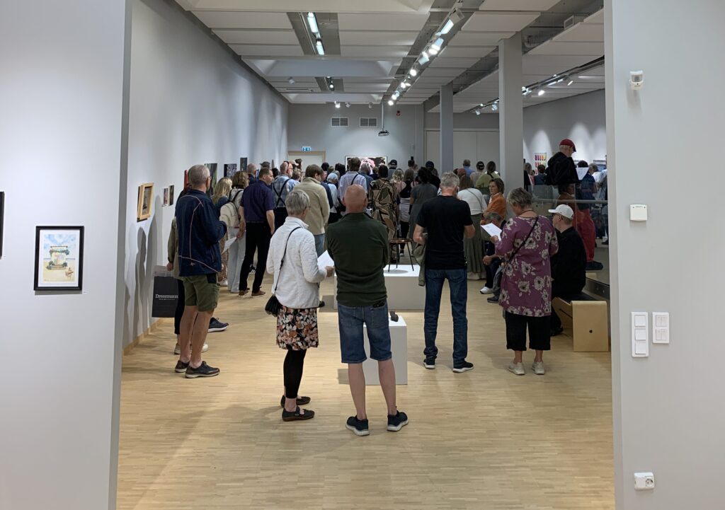Många besökare står utspridda i en stor och ljus konsthall med målningar längs väggarna. Flera personer tittar på konst eller läser programblad.