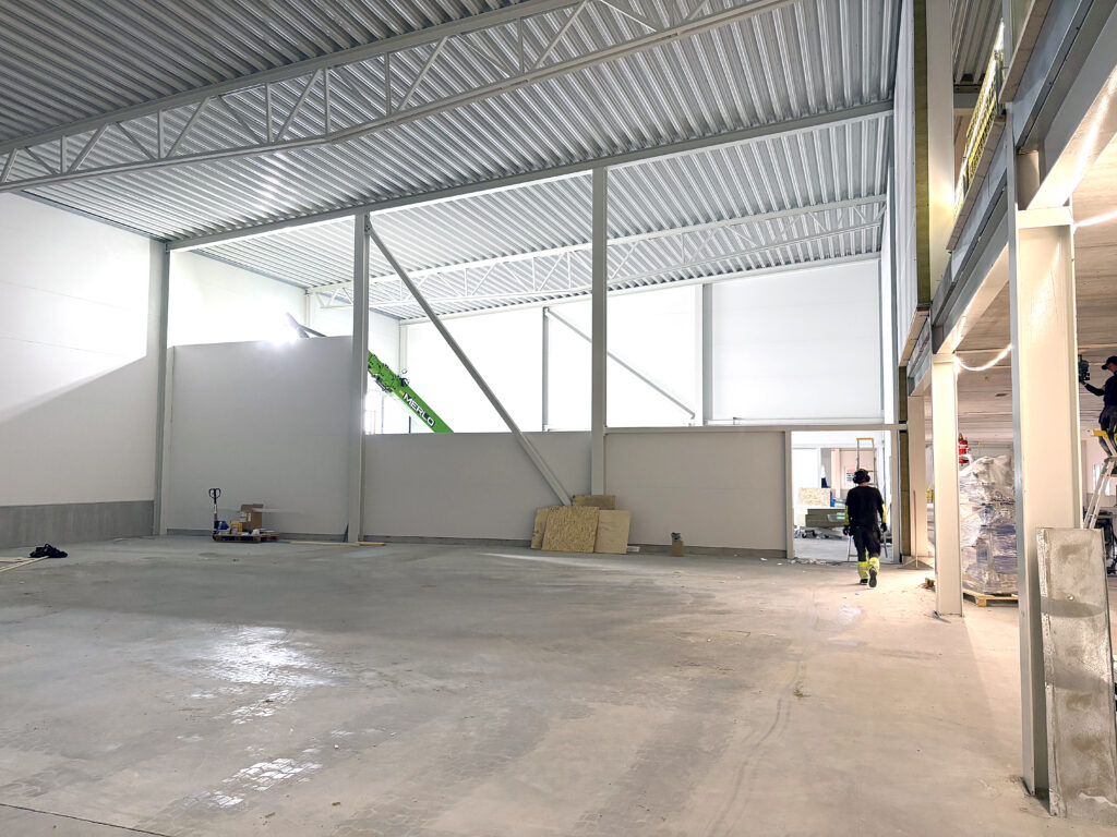 Invändigt foto av en rymlig hall med högt i tak, stålbalkar och nygjutna golv. En person går genom lokalen mot en öppning längre in.