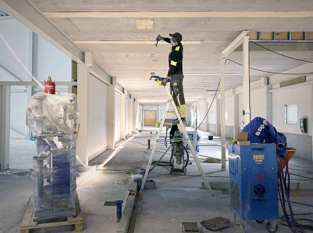 En byggarbetare står på en stege och arbetar i taket längs en bred korridor. Maskiner, verktyg och material står utplacerade runt arbetsområdet.