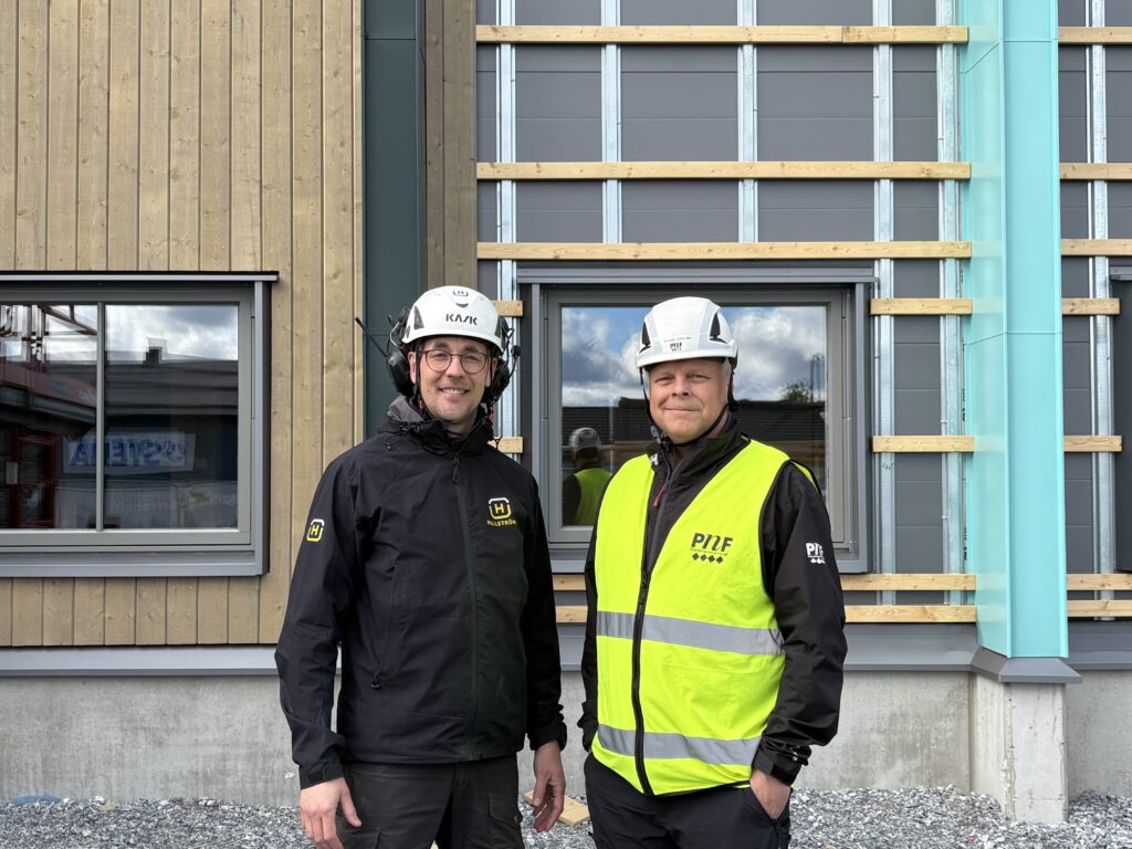 Två personer i skyddshjälmar står framför en ny industribyggnad. Bakom dem syns träfasad, fönster och färgmarkerade fasadpartier under montering.